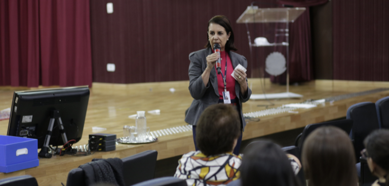 Fotografia de uma mulher, que veste calça preta, blazer cinza e camisa rosa, falando ao microfon...