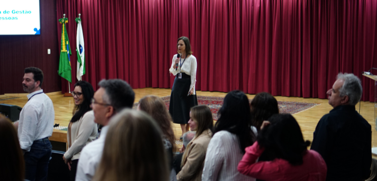 Fotografia de uma mulher, que veste camisa branca e saia preta, falando ao microfone em um palco...