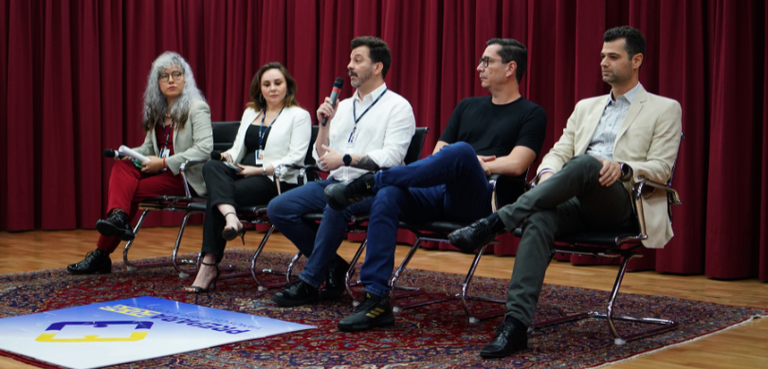 Sobre um palco e sentados em poltronas pretas com braços e pés cromados, duas mulheres e três ho...