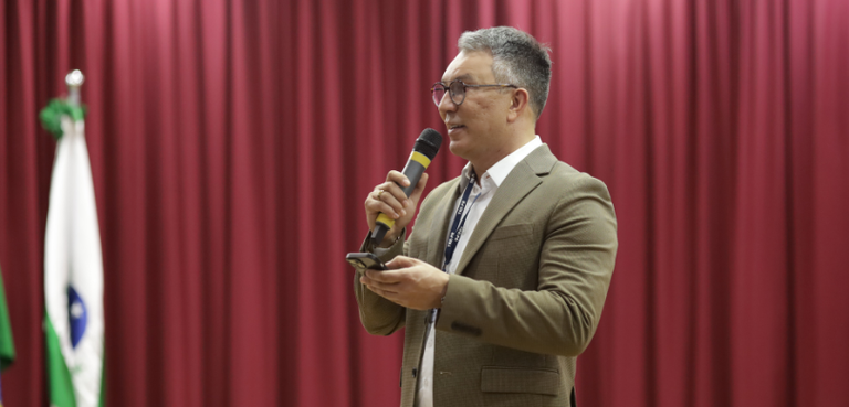 Fotografia de um homem de terno marrom falando ao microfone. Ao fundo, há uma cortina vermelha.