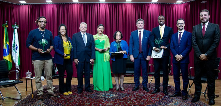 Foto de seis homens e três mulheres em pé, sobre o palco de um auditório. Alguns deles seguram u...