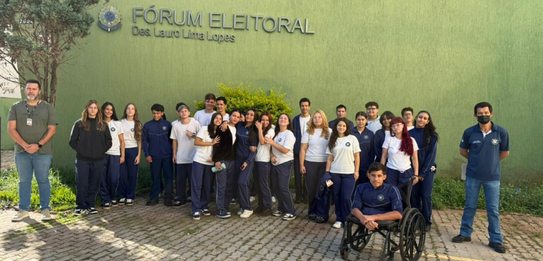 Fotografia que retrata um grupo de jovens posando para uma foto em frente à fachada do Fórum Ele...