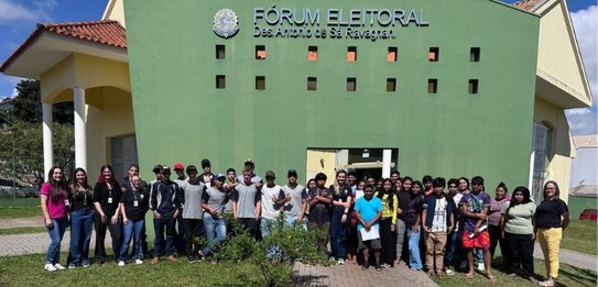 Fotografia na qual se observa um grupo de jovens posando para uma foto em frente a um prédio púb...