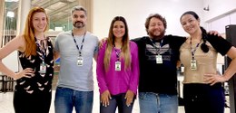 Fotografia de cinco pessoas em pé olhando para frente. Elas estão abraçadas e usam o crachá da J...