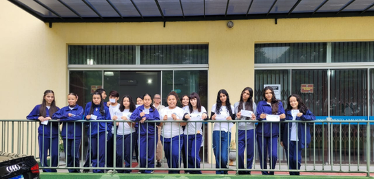 Fotografia na qual se observa um grupo de jovens posando para uma foto em frente a um prédio púb...