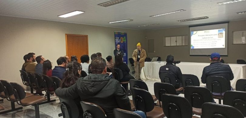Em uma sala com paredes brancas, um homem em pé discursa para em torno de quinze pessoas que est...