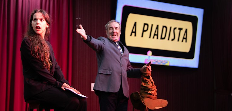 Fotografia de um auditório, em que quatro mulheres e um homem aparecem sentados em cadeiras pret...