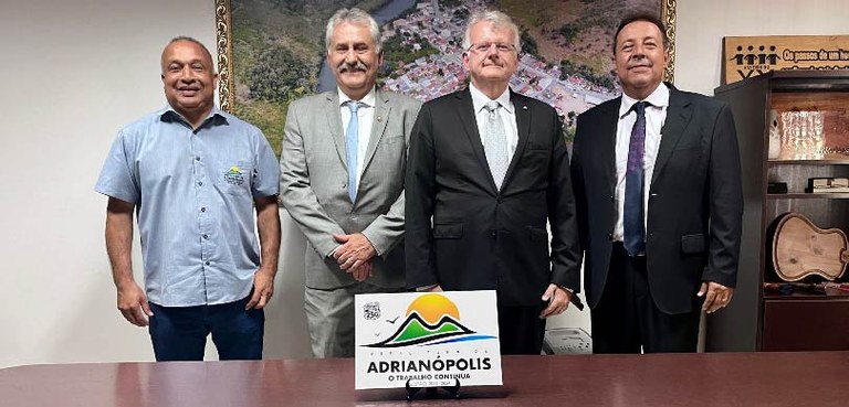 Foto de quatro homens sorrindo atrás de uma mesa de madeira. À frente deles há uma placa branca ...