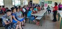 Em uma sala de aula, vários indígenas assistem sentados a uma mulher vestida com camisa vermelha...