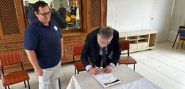 Na foto, um homem, com paletó e gravata, usando óculos, assina um documento acima de uma mesa, c...