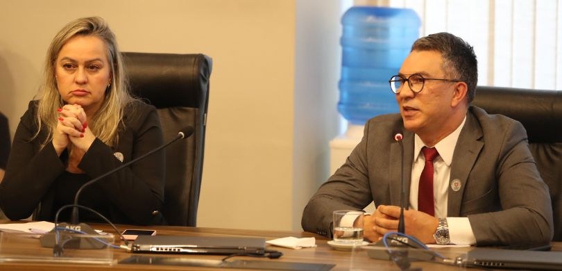 Fotografia de uma mulher e um homem sentados em cadeiras de couro e debruçados sobre uma mesa. A...