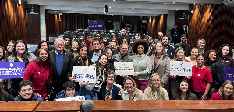 Fotografia de diversas pessoas (a maioria mulheres) posando sorrindo em uma sala com paredes de ...