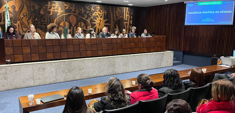 Fotografia da mesa de autoridades da audiência pública, composta por 10 mulheres e um homem. Em ...
