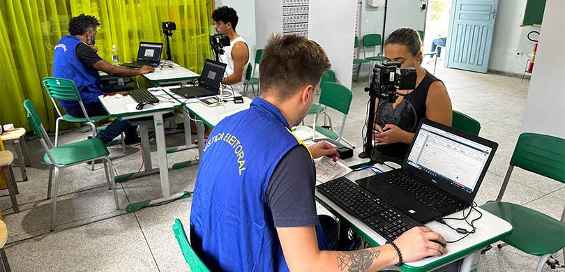 Dois homens sentados de costas com coletes azuis, onde se lê Justiça Eleitoral, estão à frente d...