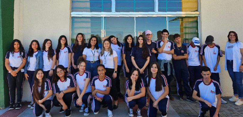 Quinze alunas, seis alunos e uma professora posam para a foto em frente a uma porta de vidro. Da...