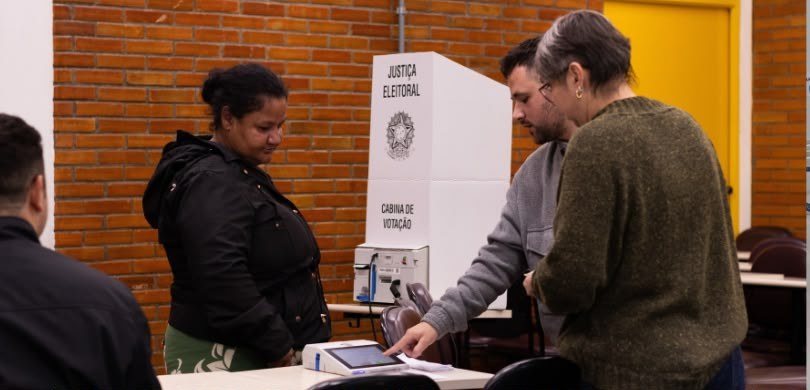 Fotografia em que se observa um ambiente interno de votação com quatro pessoas. Em primeiro plan...