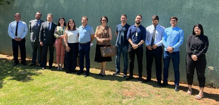 Fotografia de homens e mulheres em frente a uma parede verde. Eles estão em pé sobre um gramado.