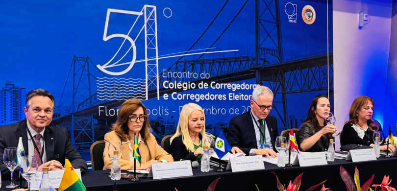 Fotografia de uma mesa de autoridades composta por quatro mulheres e dois homens. A bancada é pr...