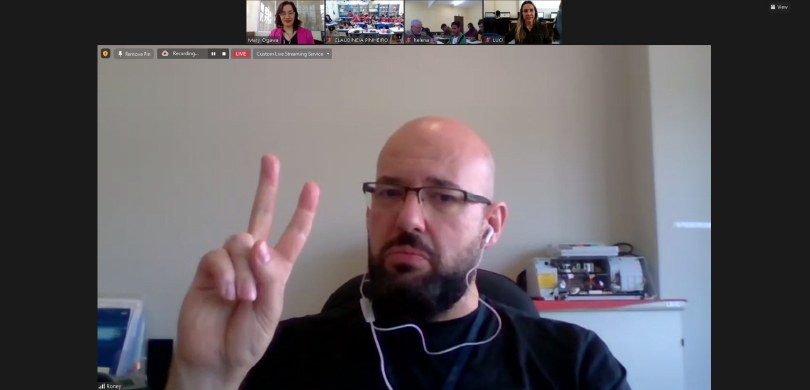 Foto de tela da reunião pela plataforma Zoom. Em primeiro plano, há um homem, de óculos, com doi...