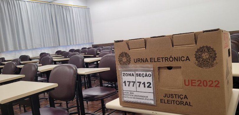 Foto em que aparece, em primeiro plano, do lado direito, uma caixa de urna sobre uma carteira es...