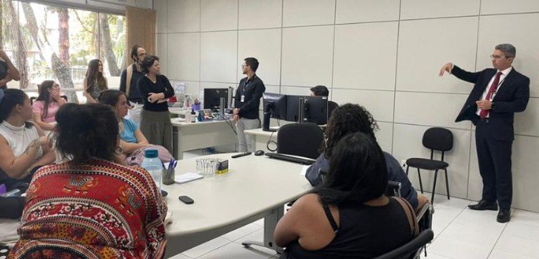 Estudantes de Pedagogia da UFPR conhecem instalações e ações do TRE-PR Fotografia de uma sala com paredes e chão branco. Do lado esquerdo, há mulheres sentadas em cade...