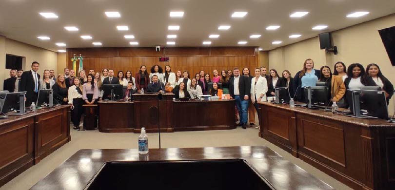 Fotografia de 13 pessoas, entre homens e mulheres, posando sorrindo na sala de sessões do TRE-PR...