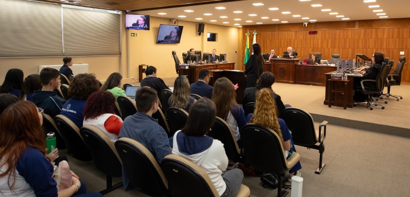 Fotografia na qual é possível ver estudantes sentados em uma platéia, olhando para a mesa da Cor...
