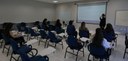 Foto de uma sala de aula com sete mulheres sentadas em cadeiras azuis com estrutura em ferro pre...
