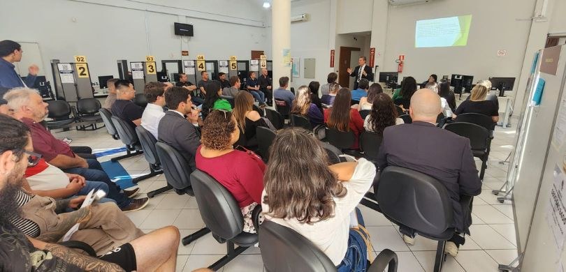 Fotografia de homens e mulheres sentados de costas em cadeiras com estofado preto. Ao fundo, pod...