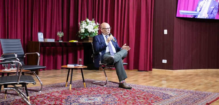 Homem, de terno e gravata, palestra sentado em uma cadeira sobre o palco de um auditório. Ao lad...