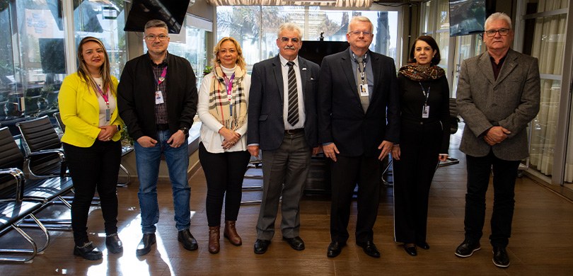 Fotografia na qual quatro homens e três mulheres estão posando em uma sala de reunião. Na sala é...