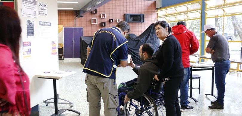 Fotografia em que se veem seis pessoas de costas em um local de votação. Quase no centro da imag...
