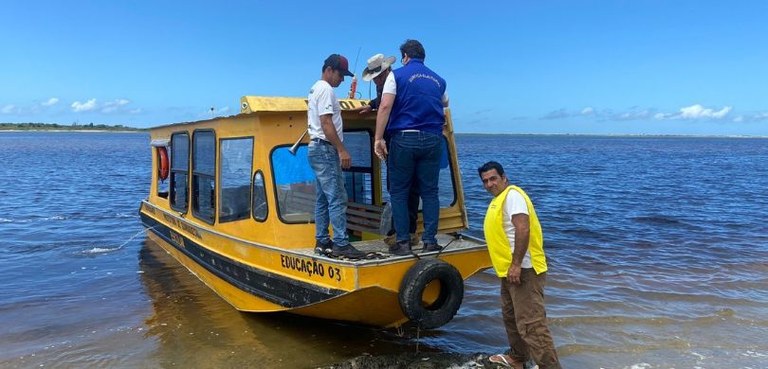 Fotografia em que se veem três pessoas na proa de um pequeno barco amarelo (um deles, de costas,...