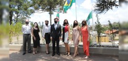 Fotografia de um grupo de pessoas em pé. Elas usam máscara e olham para frente. Atrás, há as ban...
