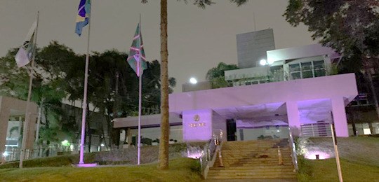 Foto tirada à noite da fachada do TRE-PR iluminada na cor roxa. Do lado esquerdo da foto, estão ...