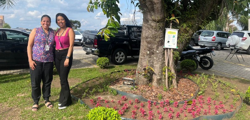 Duas mulheres posam para a foto ao lado da árvore em que está a colmeia e da placa que informa s...