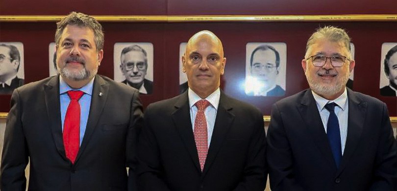 Três homens em pé, trajando ternos, posam para a foto, em frente a quadros em preto e branco.