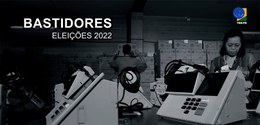 Foto em preto e branco, na qual se observam urnas eletrônicas em primeiro plano sobre uma bancad...
