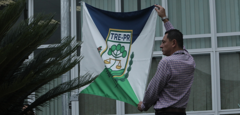 SECTI Fotografia de um homem segurando a bandeira do TRE-PR esticada. A bandeira é das cores branca, a...