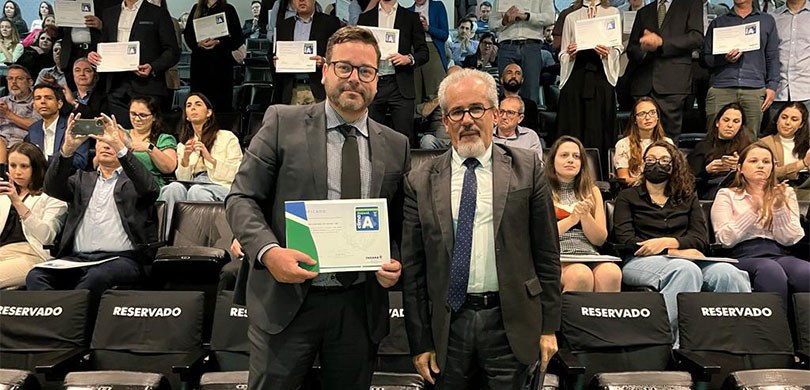 Fotografia de dois homens em pé, vestindo terno. Um deles está segurando um certificado em mãos....
