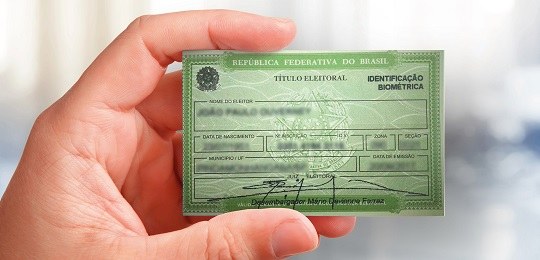 Em primeiro plano, observa-se a mão de uma pessoa segurando um título eleitoral, um documento ve...