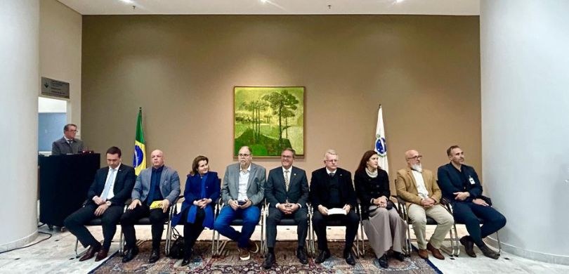 Fotografia de duas mulheres e sete homens sentados em cadeiras, sobre um tapete colorido e um pi...