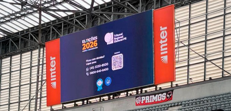 Fotografia de um grande telão digital instalado em um estádio de futebol sob uma estrutura de te...