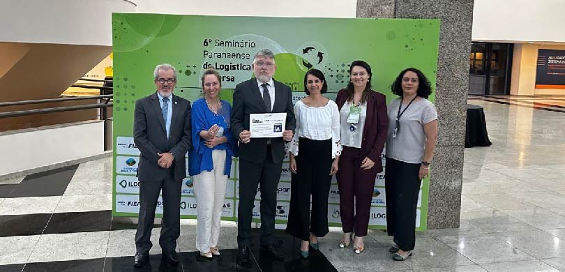 Em um grande hall de um prédio comercial, dois homens e quatro mulheres posam para a foto. Um do...