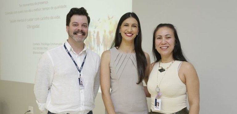 Fotografia na qual duas mulheres e um homem posam sorrindo. Ao fundo, é possível ver uma parede ...