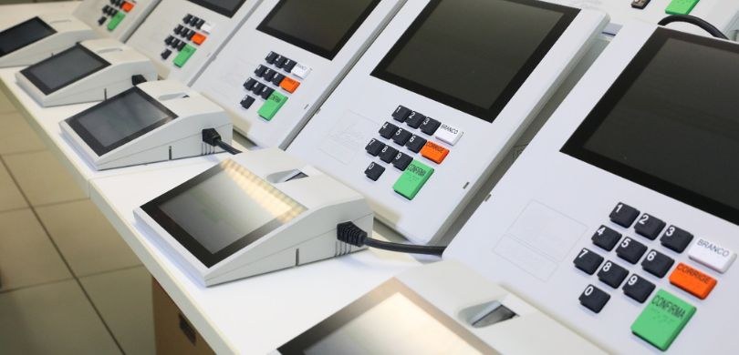 Fotografia de uma bancada branca sobre a qual se veem partes de cinco urnas eletrônicas modelo 2...