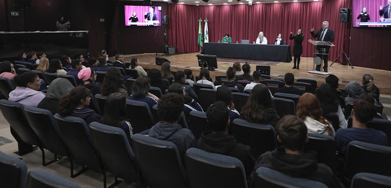Fotografia de homens e mulheres sentados em cadeiras com estofado azul-escuro de frente ao palco...